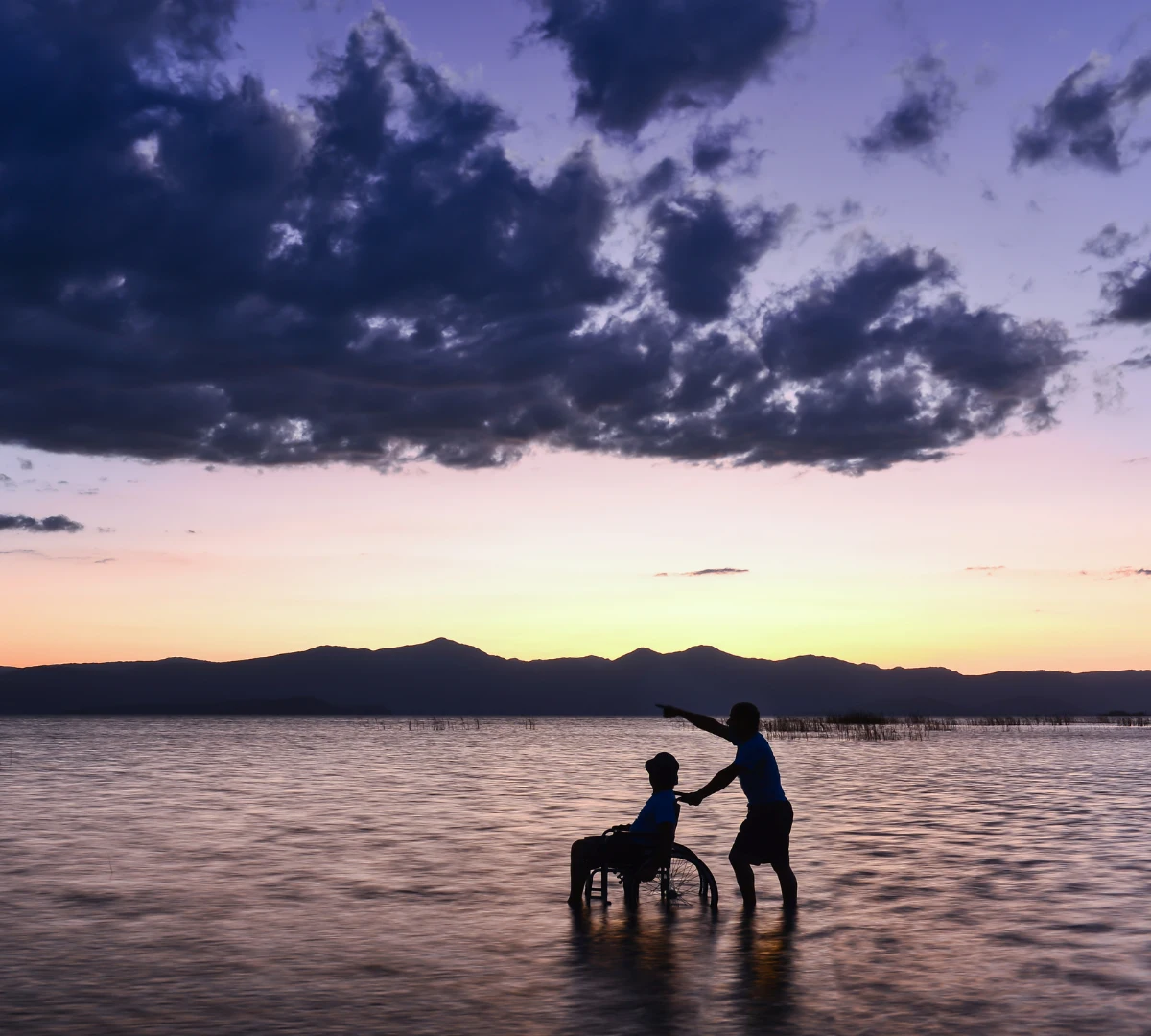 Disabled person in wheelchair in shallow water with support worker watching sunset
